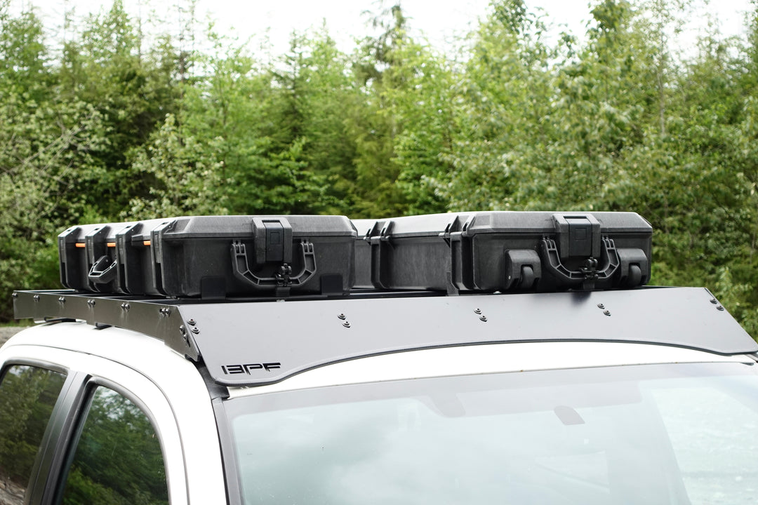 BPF Universal Pelican Vault V700 Case Mounting brackets installed on a vehicle roof rack securely holding multiple black Pelican Vault cases against a forest backdrop.