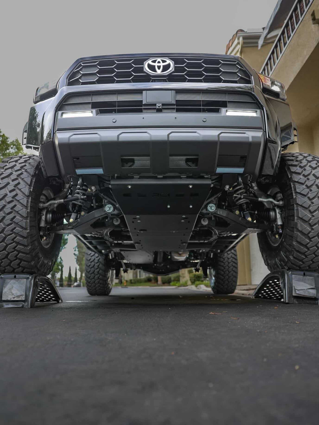 Underside view of a black Toyota truck elevated on ramps showcasing the BPF 2024+ Tacoma,4runner and Land Cruiser Tow Hooks Recovery Points mounted on the front frame with heavy-duty off-road tires and suspension components visible.