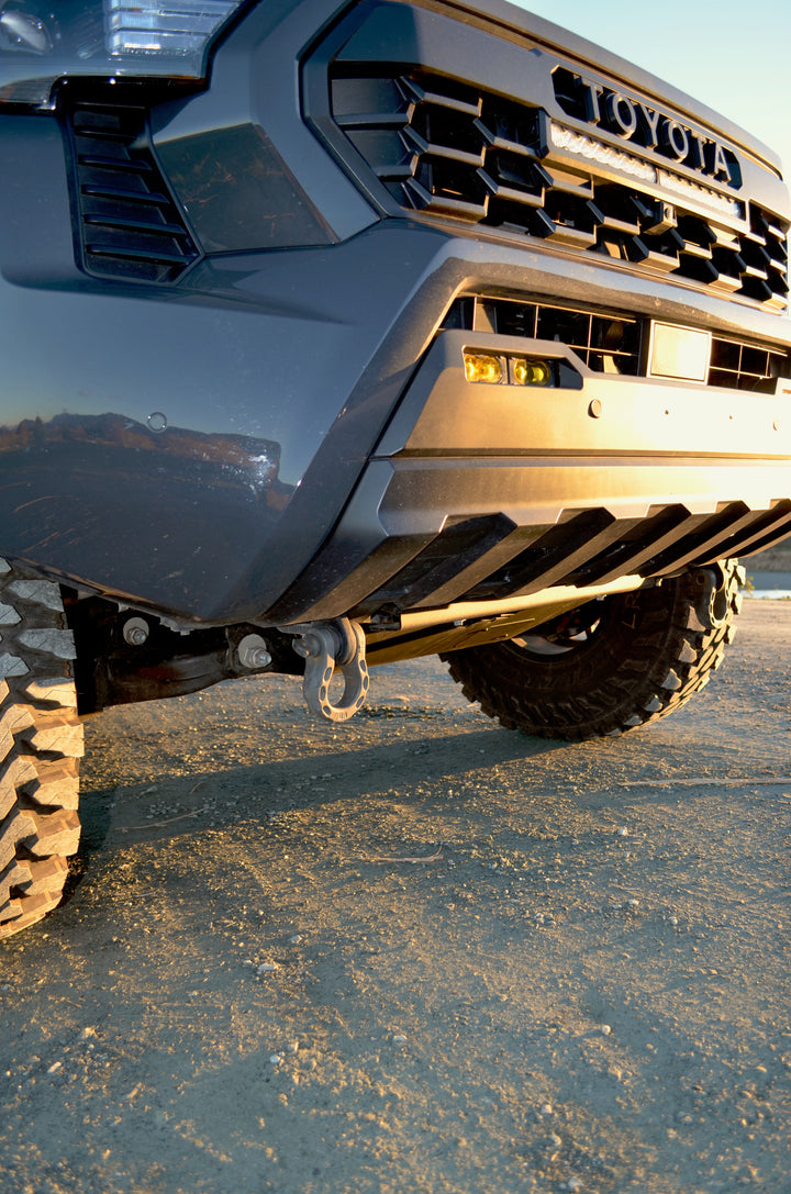 Close-up of the front underside of a Toyota truck featuring rugged off-road tires and a mounted BPF 2024+ Tacoma Recovery Points tow hook in a dirt outdoor setting at sunset.