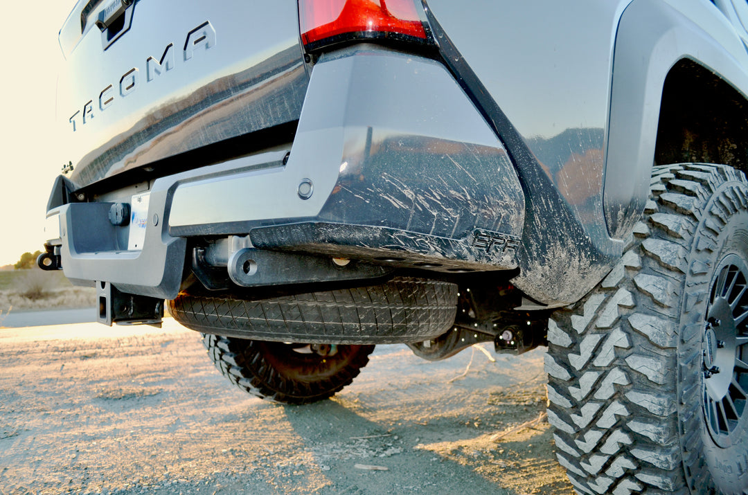 BPF 4th gen Tacoma rear tow hooks with integrated skid plates installed on a gray Tacoma truck showing heavy-duty off-road tires and rugged terrain background.