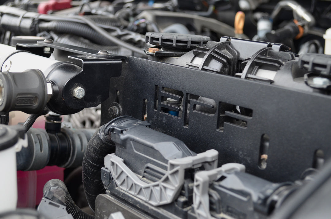 Close-up view of a CNC fabricated black metal twin compressor mount installed in an engine bay with "BPF" cutout, showcasing the BPF 2024-2025 Tacoma and Land Cruiser Twin Compressor Mount designed for durability and OEM fitment.
