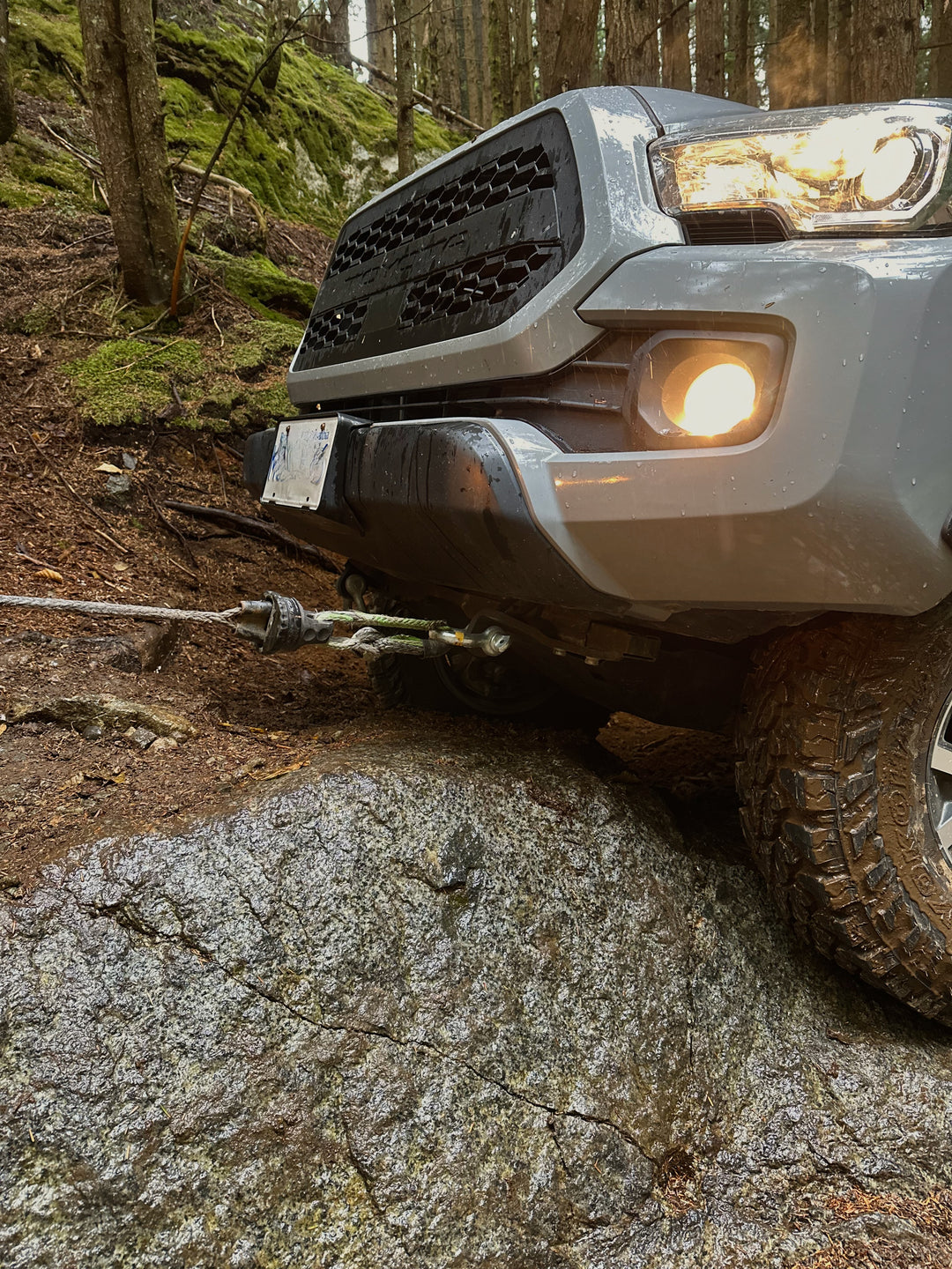 Front view of a silver off-road vehicle with a lit fog light and aggressive tire gripping a large rock, connected to a winch cable, parked on a forest trail with mossy trees.
