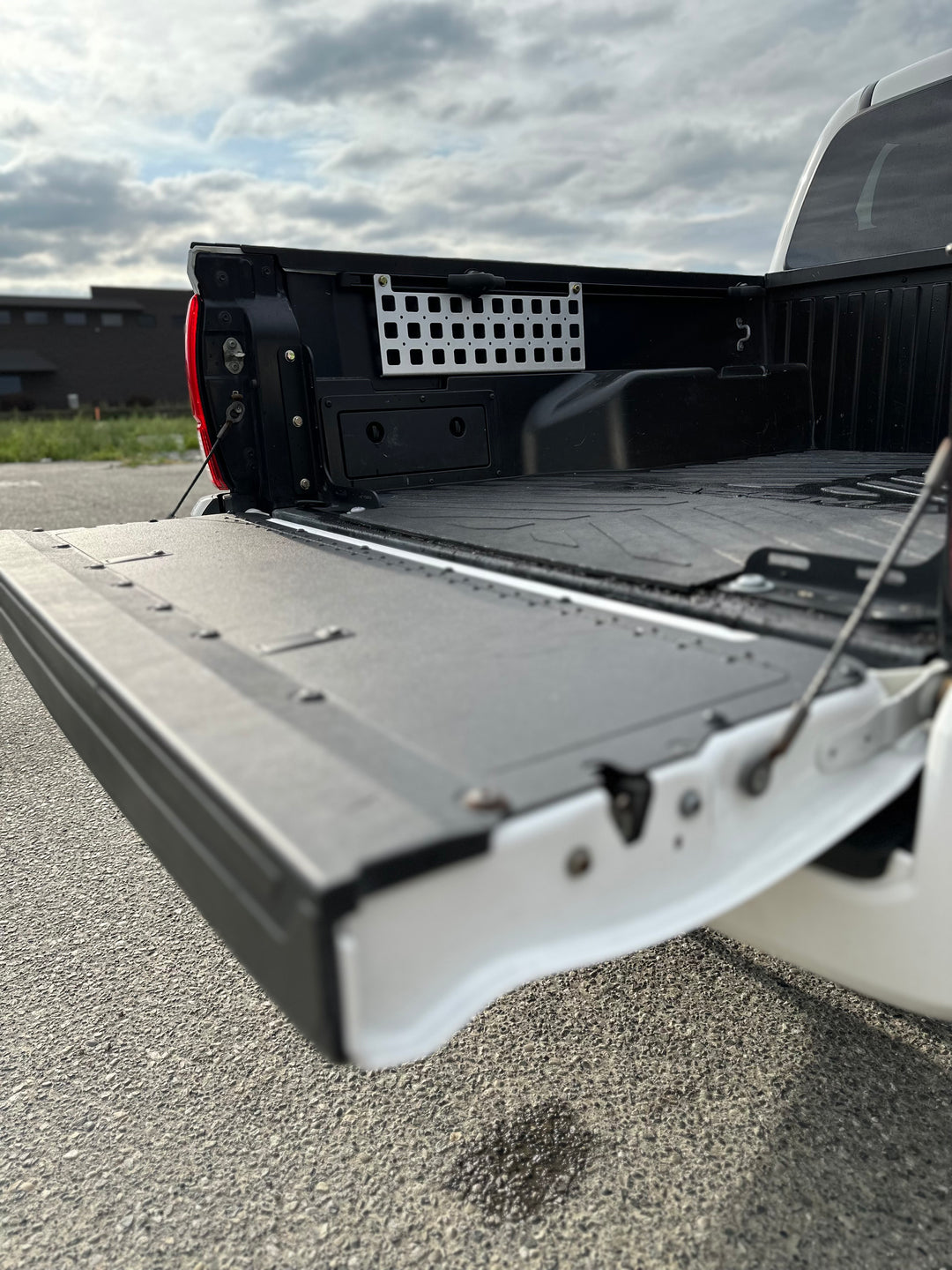 Open tailgate of white truck showing installed BPF 05-23 Tacoma bed Molle panels on the inner side of the truck bed wall under cloudy sky.
