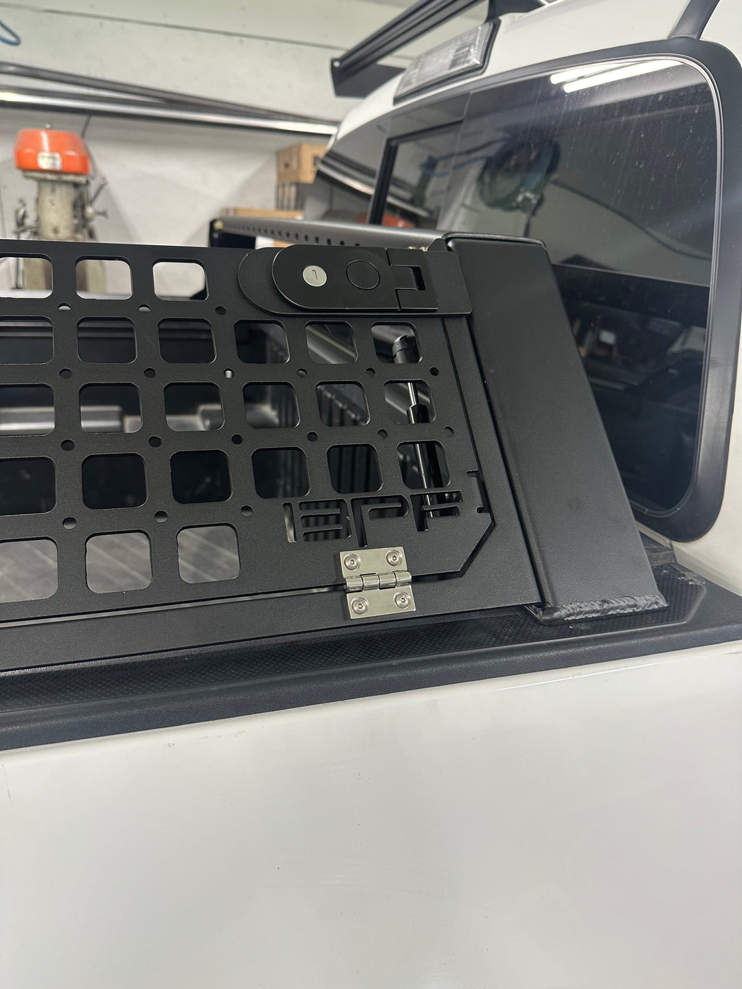 Close-up of a black metal bed rack with grid mesh and latch mechanism installed on a white truck bed, showcasing the 05-26 Tacoma Bed Rack fits 2nd,3rd and 4th gen Tacomas design and build quality.