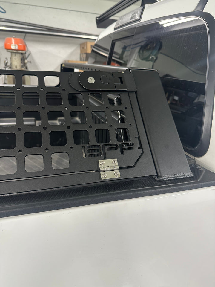 Close-up of a black metal bed rack with grid mesh and latch mechanism installed on a white truck bed, showcasing the 05-26 Tacoma Bed Rack fits 2nd,3rd and 4th gen Tacomas design and build quality.