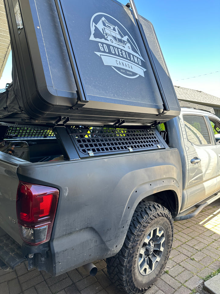 Black metal bed rack installed on a grey pickup truck with a rooftop camping tent open, featuring the logo "Go Overland Canada," showing rugged tires and off-road setup with the 05-26 Tacoma Bed Rack fits 2nd,3rd and 4th gen Tacomas.