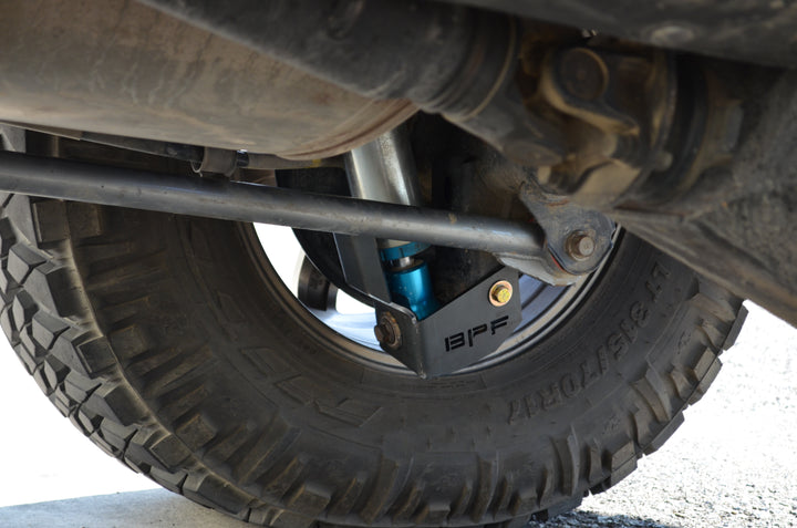 Close-up view of an off-road tire fitted with BPF 4runner 04-24/FJ 07-14 Shock Guards protecting the shock absorber, showcasing durable powder-coated metal construction and OEM fitment.