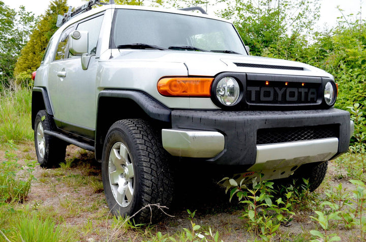 Silver SUV equipped with a BPF Completed 2007-2014 FJ Cruiser Grill featuring bold Toyota lettering, parked on grassy terrain with surrounding greenery and rugged tires.