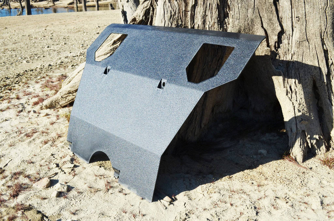 BPF 2005-2015 Toyota Tacoma Skid Plate made from durable textured metal, shown outdoors leaning against a tree on sandy ground.