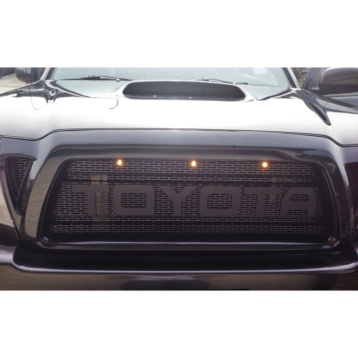 Close-up of a black truck grille featuring the BPF 2005-2011 Toyota Tacoma Raptor Style Mesh and Lettering with integrated lights and a rugged, aggressive design.