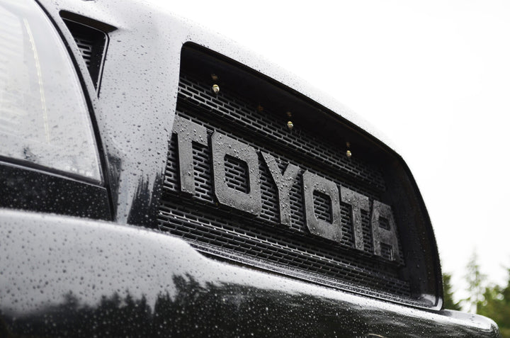 Close-up view of a black truck front grille with large bold lettering spelling TOYOTA, featuring the BPF 2005-2011 Toyota Tacoma Raptor Style Mesh and Lettering in a textured black finish with water droplets on the surface.