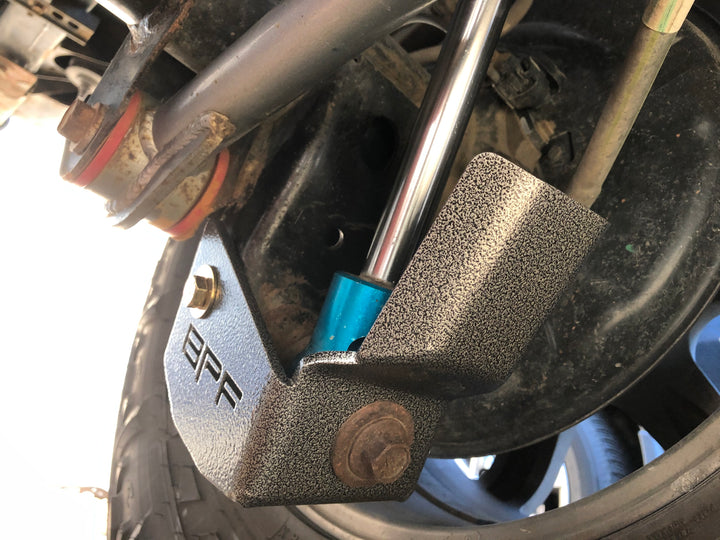 Close-up of BPF 4runner 04-24/FJ 07-14 Shock Guards installed on vehicle suspension showing textured powder-coated metal guard protecting shock absorber mount.