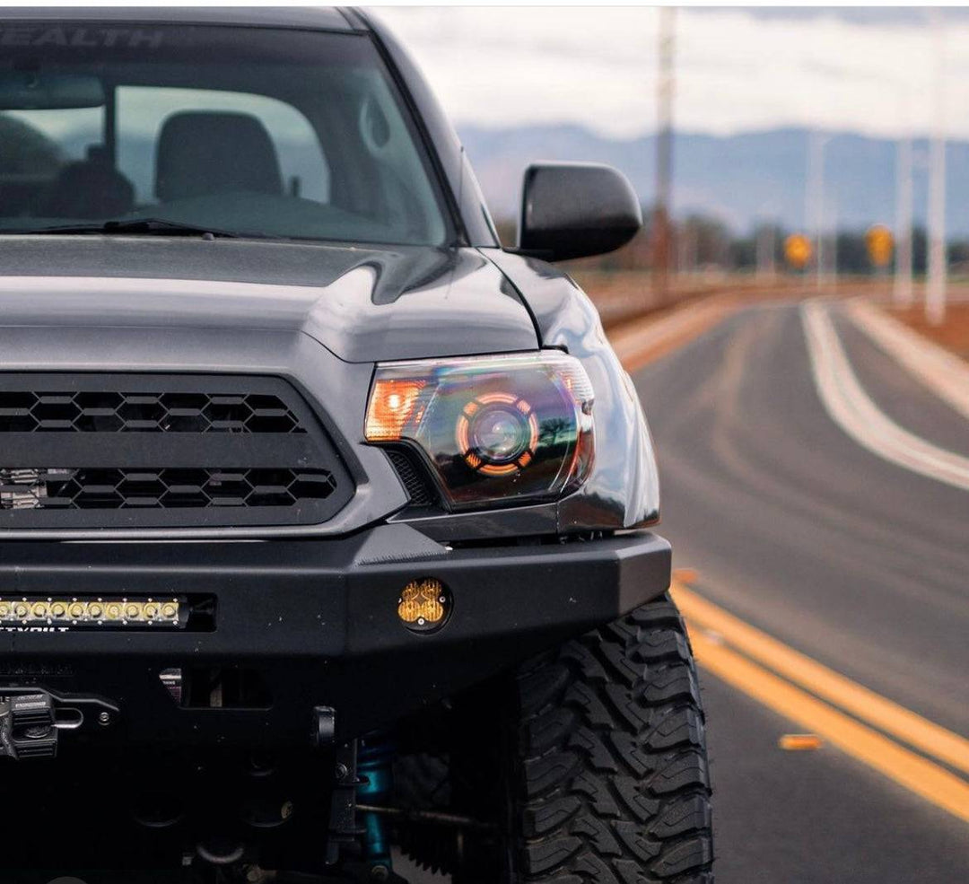 Close-up of a black Toyota Tacoma featuring the BPF 2012-2015 Toyota Tacoma Grill with rugged off-road tires and a heavy-duty front bumper on a paved road.