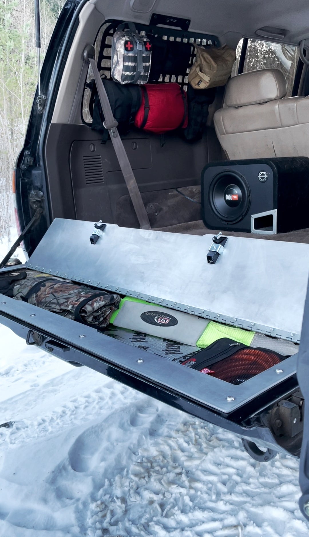 BPF 80 Series Land Cruiser and LX450 Tailgate Storage showing an open metal storage compartment on the tailgate with organized gear inside, mounted in a vehicle on a snowy surface.