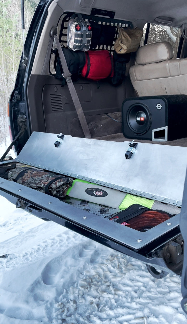 BPF 80 Series Land Cruiser and LX450 Tailgate Storage showing an open metal storage compartment on the tailgate with organized gear inside, mounted in a vehicle on a snowy surface.
