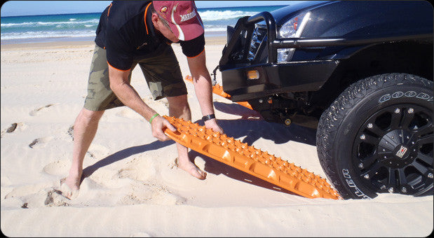 Man using bright orange MAXTRAX MKII traction boards to help a black off-road vehicle stuck in deep sand on a beach with ocean waves in the background