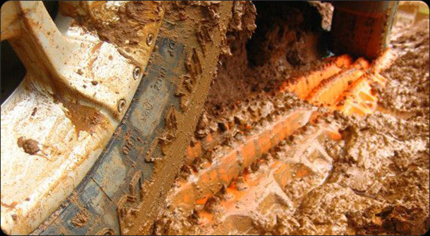 MAXTRAX MKII recovery traction boards partially buried in thick mud next to a muddy tire, designed for off-road vehicle traction and recovery in challenging terrain.