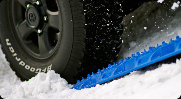MAXTRAX MKII traction board in bright blue positioned under a vehicle tire on snow, designed to provide grip and recovery assistance in off-road conditions.